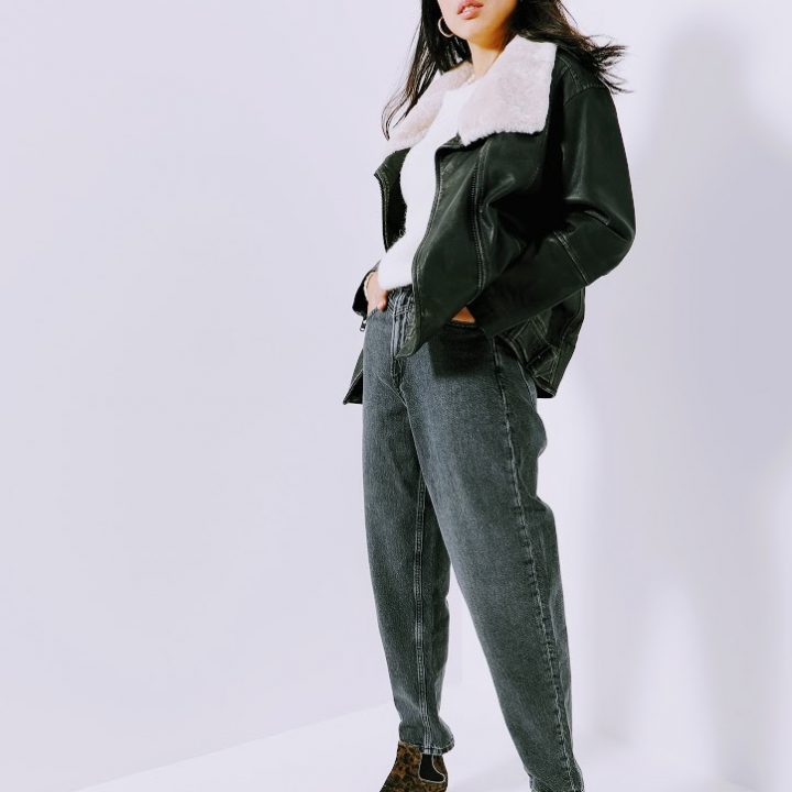 Eine Frau posiert selbstbewusst. Sie trägt eine schwarze Lederjacke mit weißem Pelzkragen, graue Jeans und Stiefel mit Leopardenmuster. Der Hintergrund ist eine schlichte weiße Wand, die den Kontrast zu ihrem Outfit verstärkt.