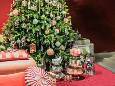 Ein wunderschön geschmückter Weihnachtsbaum mit Ornamenten steht neben einem Tisch mit Teetassen, einem mehrstöckigen Tablett mit Kerzen und einer Kaffeemaschine. Daneben liegen verpackte Geschenke auf einem roten Teppich und sorgen für eine festliche Weihnachtsszene.