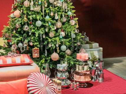 Ein wunderschön geschmückter Weihnachtsbaum mit Ornamenten steht neben einem Tisch mit Teetassen, einem mehrstöckigen Tablett mit Kerzen und einer Kaffeemaschine. Daneben liegen verpackte Geschenke auf einem roten Teppich und sorgen für eine festliche Weihnachtsszene.