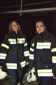 Zwei Feuerwehrmänner in dunklen Uniformen mit reflektierenden Streifen stehen drinnen. Jeder trägt einen weißen Helm. Sie lächeln in die Kamera. Der Hintergrund ist dunkel, was auf einen Innenraum oder eine schwach beleuchtete Umgebung schließen lässt.