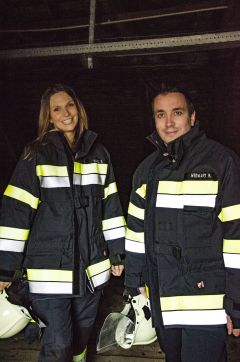 Zwei Feuerwehrmänner in dunklen Uniformen mit reflektierenden Streifen stehen drinnen. Jeder trägt einen weißen Helm. Sie lächeln in die Kamera. Der Hintergrund ist dunkel, was auf einen Innenraum oder eine schwach beleuchtete Umgebung schließen lässt.
