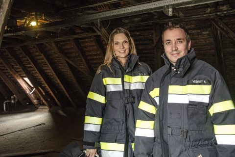 Zwei Personen in Feuerwehrausrüstung stehen auf einem Dachboden mit Holzbalken. Sie tragen beide dunkle Jacken mit Reflektorstreifen und ein sanftes Licht erhellt den Raum.