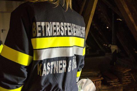 Eine Person, die eine Warnweste mit der Aufschrift „BETRIEBSFEUERWEHR KASTNER & ÖHLER“ auf dem Rücken trägt, steht in einem schwach beleuchteten Raum.