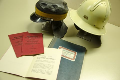 Zu einer Sammlung historischer Feuerwehrgegenstände gehören zwei Helme, einer schwarz und einer weiß, neben zwei aufgeschlagenen Heften und einer blauen Dokumentenmappe mit der Aufschrift „Niederschrift“. Die Gegenstände liegen auf einer beigen Fläche.