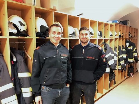 Zwei Personen in Feuerwehruniformen stehen lächelnd vor einer Wand mit Ablagefächern und halten Helme und Feuerwehrausrüstung in den Händen. Der Raum ist gut beleuchtet und beide Personen wirken stolz und selbstbewusst.