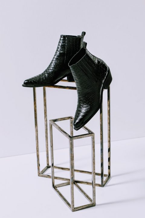 Schwarze Stiefeletten mit spitzer Spitze und strukturiertem Lederfinish werden kreativ auf zwei kubischen Metallständern vor einem hellen, neutralen Hintergrund präsentiert.