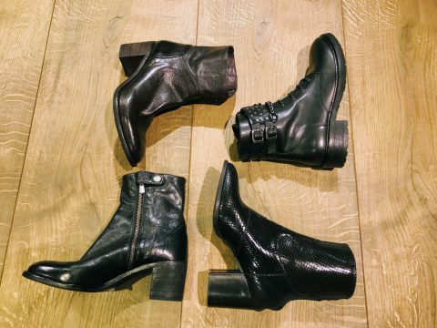 Vier schwarze Lederstiefeletten mit unterschiedlichen Absatzhöhen und Stilen sind auf einem Holzboden angeordnet. Jeder Stiefel weist unterschiedliche Details auf, wie Reißverschlüsse, Schnallen oder ein strukturiertes Muster.