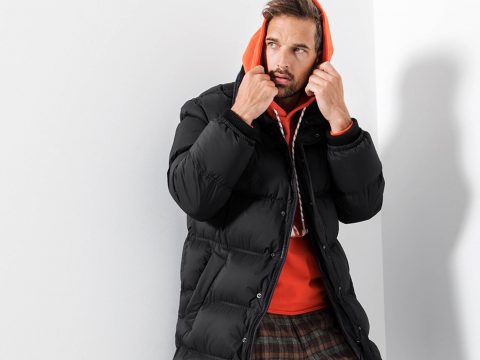 Eine Person, die einen orangefarbenen Kapuzenpullover unter einer schwarzen Pufferjacke trägt, rückt ihre Kapuze zurecht. Sie steht vor einer weißen Wand und blickt zur Seite. Sie trägt außerdem karierte Hosen.