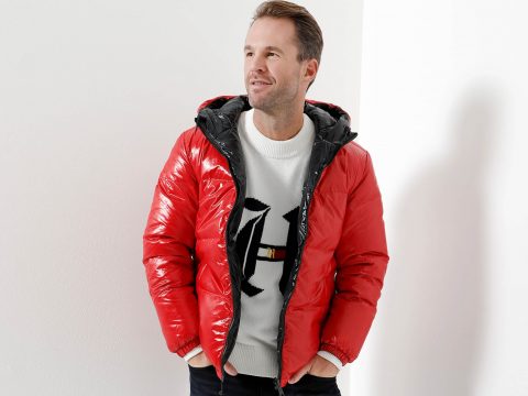 Ein Mann steht vor einer weißen Wand. Er trägt eine glänzende rote Pufferjacke über einem weißen Pullover mit einem schwarzen Buchstabenmuster. Er hat kurze Haare und einen entspannten Gesichtsausdruck. Seine Hände stecken in den Jackentaschen.