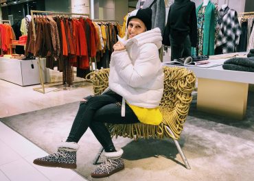 Eine Person in einer weißen Pufferjacke, einer schwarzen Beanie-Mütze und Stiefeln mit Leopardenmuster sitzt auf einem einzigartigen Strickstuhl in einem Bekleidungsgeschäft, umgeben von Kleiderständern mit bunter Kleidung.