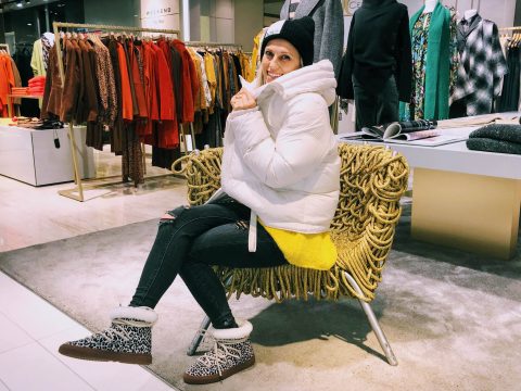 Eine Person in einer weißen Pufferjacke, einer schwarzen Beanie-Mütze und Stiefeln mit Leopardenmuster sitzt auf einem einzigartigen Strickstuhl in einem Bekleidungsgeschäft, umgeben von Kleiderständern mit bunter Kleidung.