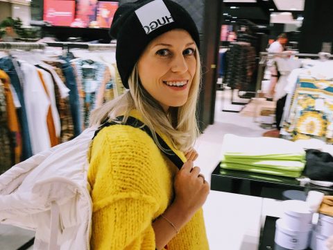 Eine Frau in einem leuchtend gelben Pullover und einer schwarzen Mütze lächelt beim Einkaufen in einem Bekleidungsgeschäft. Sie trägt eine weiße Jacke über der Schulter, im Hintergrund sind Kleiderständer und Auslagen zu sehen.