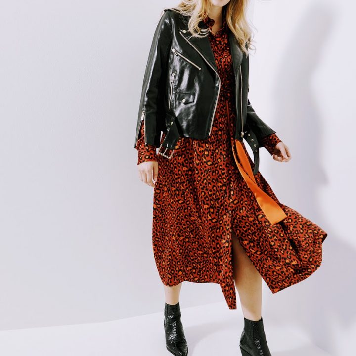 Eine Person trägt eine schwarze Lederjacke über einem rot-schwarzen Kleid mit Leopardenmuster und dazu schwarze Stiefeletten. Die Person hat lange blonde Haare und ist in Bewegung vor einem schlichten weißen Hintergrund aufgenommen.