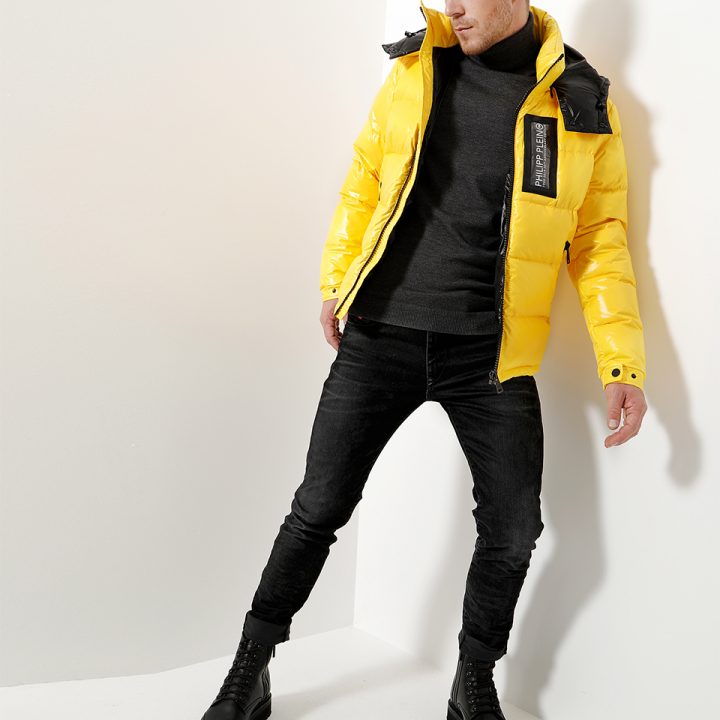 Ein Model trägt eine leuchtend gelbe Pufferjacke über einem schwarzen Rollkragenpullover und schwarzen Jeans und lehnt mit einem Bein nach vorne an einer weißen Wand. Schwarze Stiefel vervollständigen das stylische Outfit.
