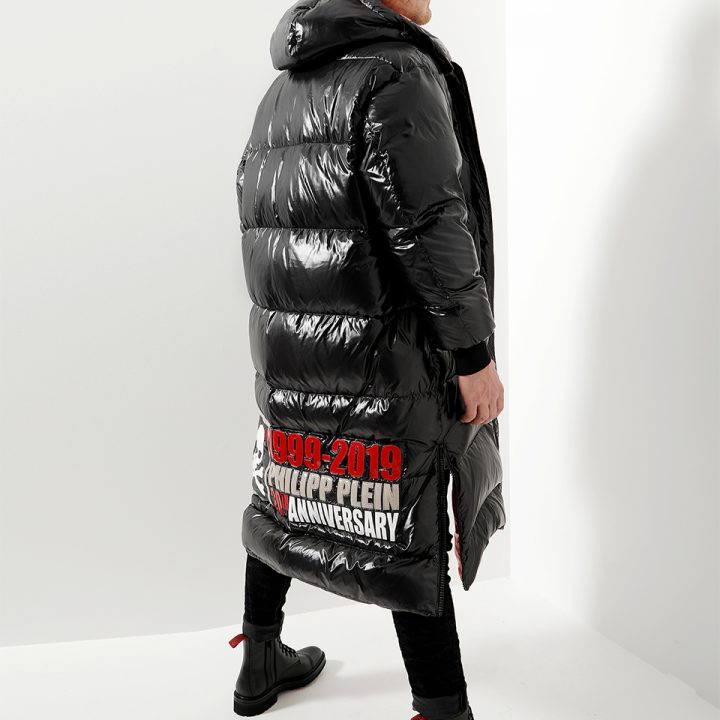 Mann trägt einen langen, glänzenden, schwarzen Puffermantel mit roter Textgrafik auf dem unteren Rücken, auf der steht „1999-2019 PHILIPP PLEIN ANNIVERSARY“. Er steht seitlich in einem weißen, minimalistischen Raum und trägt schwarze Hosen und Stiefel.