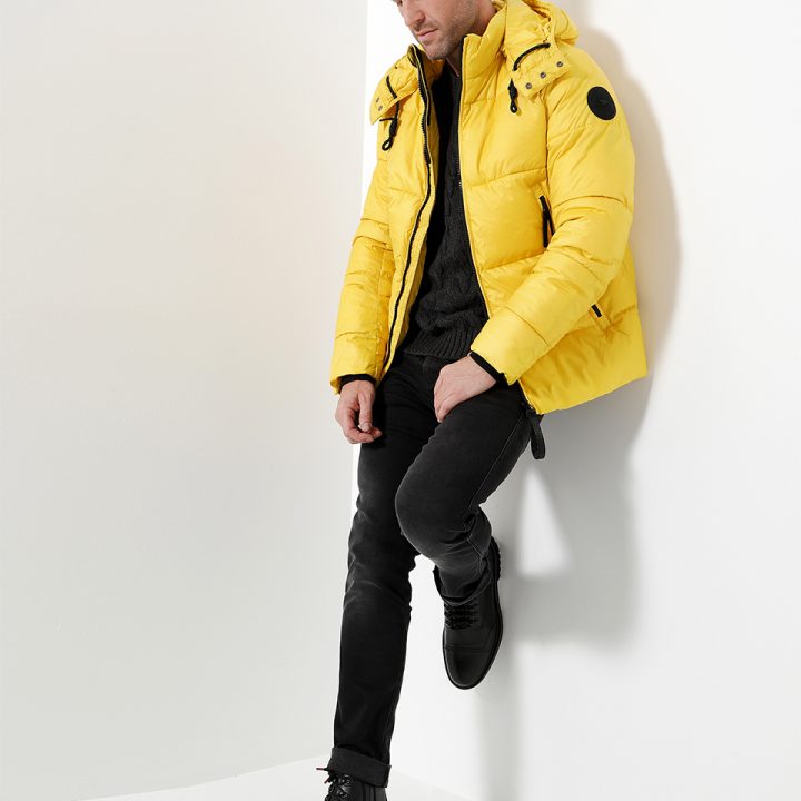 Eine Person in einer leuchtend gelben Pufferjacke, schwarzen Hosen und Stiefeln lehnt an einer weißen Wand. Die Jacke hat eine Kapuze und einen runden Aufnäher am Ärmel. Die Kulisse ist minimalistisch mit sanfter Beleuchtung.