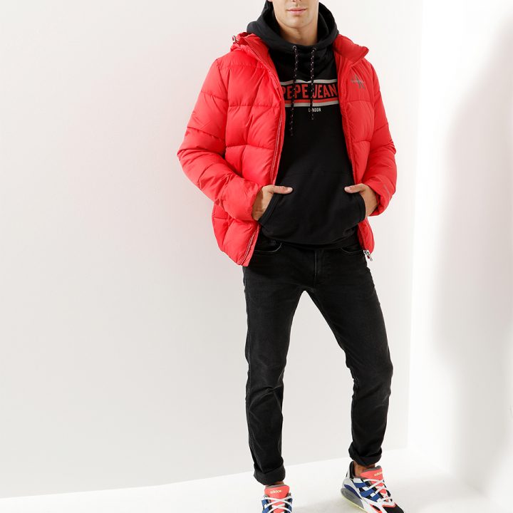 Eine Person trägt eine leuchtend rote Pufferjacke über einem schwarzen Kapuzenpullover mit der Aufschrift „Pepe Jeans“. Sie trägt außerdem schwarze Jeans und bunte Turnschuhe. Der Hintergrund ist schlicht weiß.