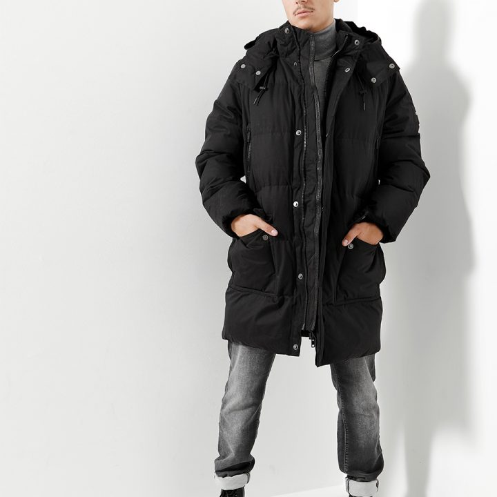 Eine Person steht vor einem weißen Hintergrund und trägt einen langen schwarzen Puffermantel mit Kapuze, einen grauen Rollkragenpullover, graue Jeans und schwarze Stiefel. Ihre Hände stecken in den Manteltaschen und sie blickt zur Seite.