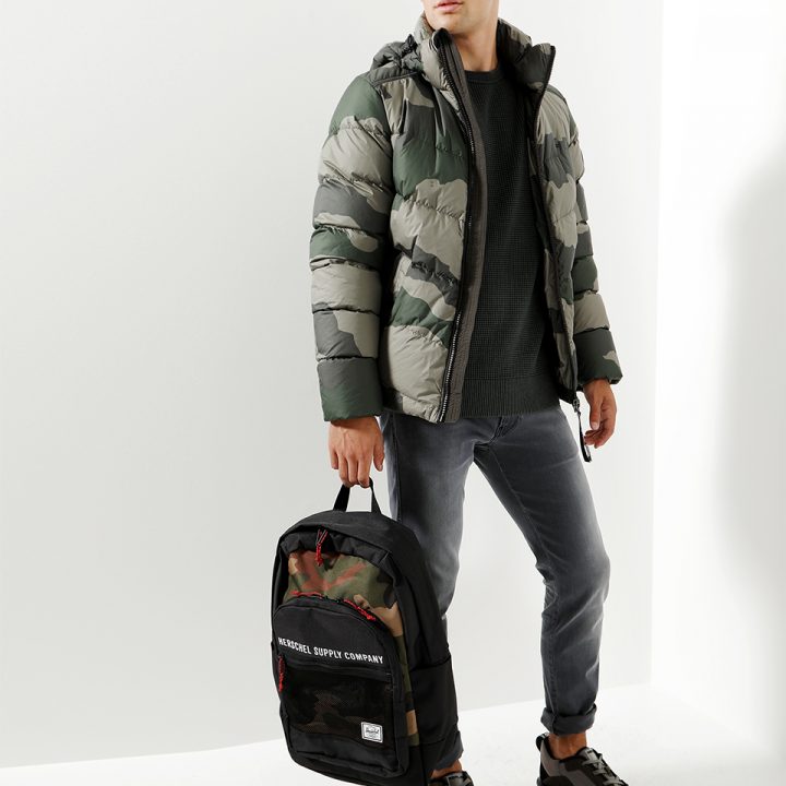 Eine Person in einer grün-grauen Pufferjacke, einem dunklen Pullover, grauen Jeans und Turnschuhen steht vor einem weißen Hintergrund. Sie hält einen Rucksack in Schwarz und Camouflage.