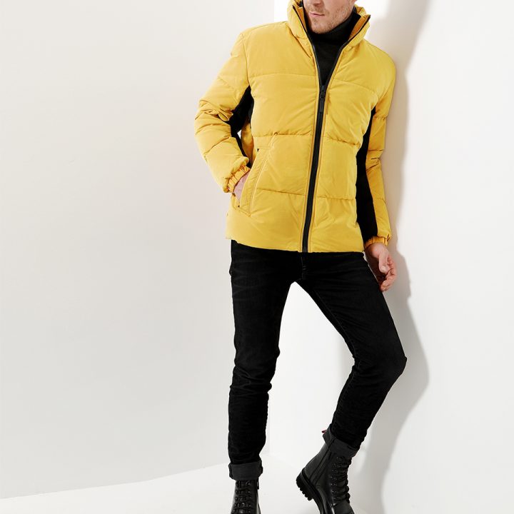 Eine Person steht vor einer weißen Wand. Sie trägt eine leuchtend gelbe Pufferjacke, einen schwarzen Rollkragenpullover, schwarze Röhrenhosen und schwarze Stiefel. Sie hat kurze Haare und schaut leicht zur Seite. Der Look ist lässig und modisch.