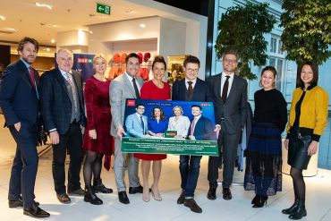 Eine Gruppe von neun Personen in formeller Kleidung steht in einem Innenbereich und hält ein großes Plakat in der Hand. Das Plakat zeigt Bilder von drei Personen und Logos. Sie befinden sich in einem hell erleuchteten Bereich, möglicherweise einem Einkaufszentrum oder Veranstaltungsort.