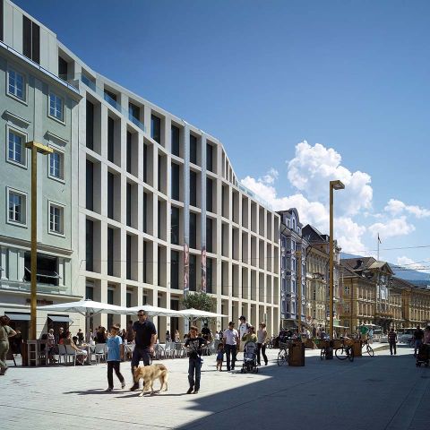 Eine moderne Straßenszene mit Passanten und einem Hund vor einem modernen mehrstöckigen Gebäude mit großen Fenstern. In der Nähe stehen Tische im Freien mit Sonnenschirmen und eine Reihe Fahrräder. Im Hintergrund ist traditionelle Architektur sichtbar.