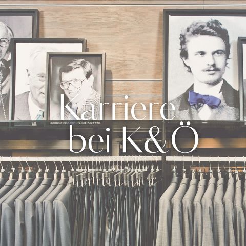 Ein Schaufenster in einem Bekleidungsgeschäft mit ordentlich aufgereihten Anzügen auf Kleiderbügeln. Über den Anzügen hängen gerahmte Schwarzweißfotos von Männern und der Text auf Deutsch lautet „Karriere bei K&O“.