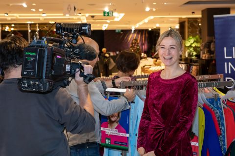 Eine lächelnde Frau in einem roten Kleid steht neben einem Kleiderständer in einem Geschäft. Ein Kamerateam filmt sie. Die Szene ist hell, im Hintergrund sind verschiedene bunte Kleidungsstücke zu sehen.