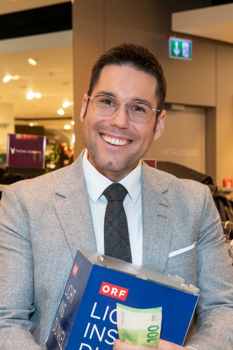 Eine lächelnde Person in hellgrauem Anzug und Brille hält ein Buch und einen 200-Euro-Schein. Im Hintergrund ist eine Innenkulisse mit Schildern und Lichtern zu sehen.