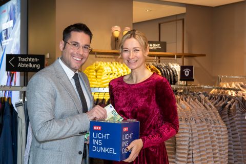 Ein Mann und eine Frau stehen lächelnd in einem Bekleidungsgeschäft. Die Frau hält eine Spendenbox mit der Aufschrift „Licht ins Dunkel“, der Mann wirft Geld hinein. Dahinter reihen sich ordentlich Kleider auf.