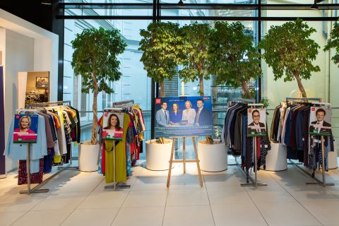 Schaufenster eines Bekleidungsgeschäfts mit Regalen voller bunter Kleidungsstücke auf beiden Seiten. In der Mitte hängt eine Staffelei mit einem großen Poster mit Porträts verschiedener Personen. Topfbäume rahmen das Schaufenster ein und helle Beleuchtung wertet das moderne Interieur auf.