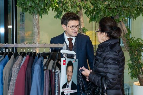 Ein Mann im Anzug steht neben einem Kleiderständer mit Jacken und spricht mit einer Frau in schwarzer Jacke und Brille. Hinter ihnen ist Grün. Auf dem Ständer ist ein Wahlplakat mit dem Gesicht eines Mannes zu sehen.