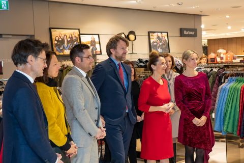 Eine Gruppe von Menschen steht in formeller Kleidung in einem Bekleidungsgeschäft. Sie stehen in einer Reihe und blicken auf jemanden außerhalb des Bildes. Im Hintergrund sind Mäntel und Jacken ausgestellt.