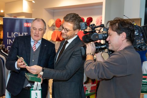 Zwei Männer in Geschäftskleidung stehen in einem Geschäft, halten gemeinsam einen Artikel in der Hand und untersuchen ihn. Ein Kameramann mit einer Videokamera filmt sie. Im Hintergrund sind bunte Hüte und Kleidungsstücke zu sehen.