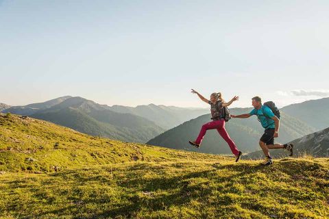 Ein Mann und eine Frau wandern fröhlich auf einem grasbewachsenen Bergkamm, mit Rucksäcken und heller Kleidung. Die sonnige Landschaft bietet sanfte Hügel und ferne Berge unter einem klaren blauen Himmel. Die Frau springt in die Luft und der Mann streckt die Hand nach ihr aus.