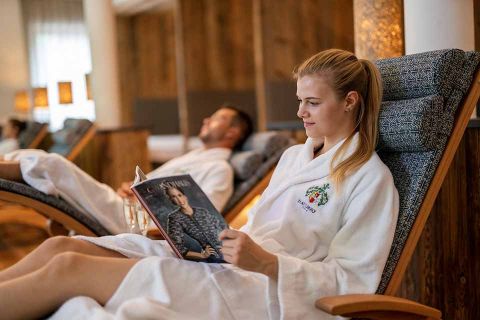 Eine Frau im weißen Bademantel liest eine Zeitschrift, während sie auf einem gepolsterten Stuhl in einem Spa liegt. Ein Mann im Hintergrund entspannt auf einem ähnlichen Stuhl. Das Zimmer ist in warmen Holztönen gehalten.