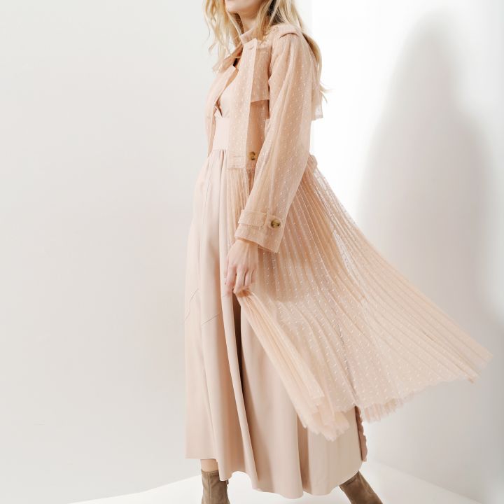 Eine Frau steht in einem hell erleuchteten Raum. Sie trägt ein fließendes pfirsichfarbenes Kleid mit einem transparenten, plissierten Überwurf. Sie hat helles Haar und blickt zur Seite. Sie trägt Stiefeletten, die zu ihrem Outfit passen.