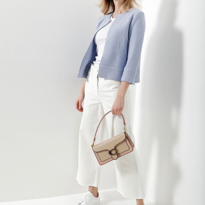 Eine Frau steht vor einer weißen Wand. Sie trägt eine hellblaue Jacke, ein weißes Oberteil und weiße Hosen mit weitem Bein. Sie hält eine beige Handtasche in der Hand und trägt weiße Turnschuhe. Ihr Haar weht leicht und sie blickt in die Kamera.