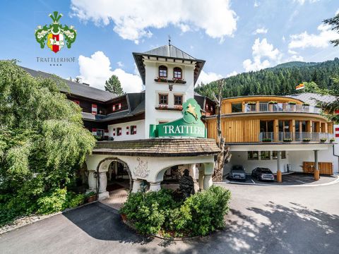 Ein malerischer Hoteleingang mit turmartiger Struktur und viel Grün. Das Gebäude verfügt über Holzakzente und einen gewölbten Steineingang. In der oberen linken Ecke erscheint ein Logo mit einem grünen Pferd und einem Schild. Im Hintergrund sind Berge und Bäume zu sehen.