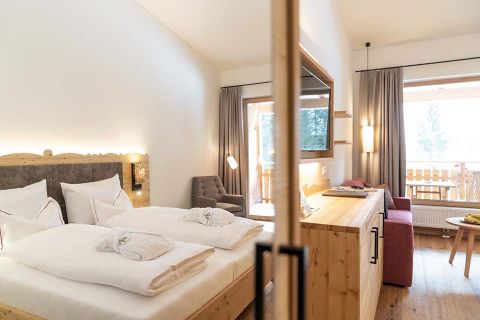 Ein gemütliches Hotelzimmer mit einem großen Bett mit weißer Bettwäsche, Holzmöbeln und einem Fernseher an der gegenüberliegenden Wand. Es gibt einen Sitzbereich mit einem roten Sofa, einem Stuhl und einem Holztisch. Durch das Fenster ist ein Balkon mit einem Holzgeländer sichtbar.