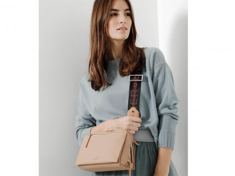 Eine Person mit langen braunen Haaren trägt einen hellblauen Pullover und einen blaugrünen Rock und hält eine beige Umhängetasche mit breitem Riemen über der Schulter. Der neutrale Hintergrund unterstreicht den lässigen Modestil.