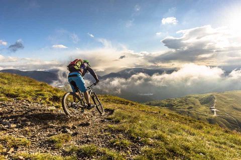Ein Radfahrer mit Rucksack fährt einen felsigen Bergpfad bergab. Die Sonne scheint durch vereinzelte Wolken und beleuchtet die üppig grüne Landschaft und die Berge in der Ferne.