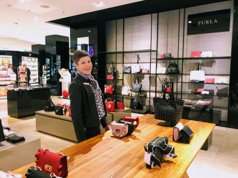 Eine Person steht in einem Geschäft und stellt verschiedene Handtaschen aus. Im Hintergrund sind Regale voller Handtaschen zu sehen und auf einem Schild steht „Furla“. Die Person lächelt und trägt einen Schal und eine dunkle Jacke.