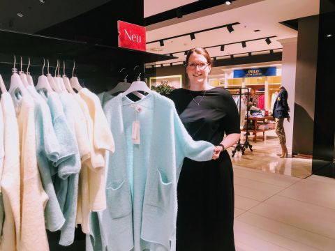 Eine Frau in einem schwarzen Kleid steht in einem Bekleidungsgeschäft und hält einen hellblauen Pullover von einer Kleiderstange. Um sie herum sind weitere Kleidungsstücke ausgestellt. Der Laden ist hell beleuchtet und ein Schild mit der Aufschrift „Neu“ ist zu sehen.