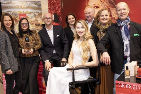 Eine Gruppe von Menschen posiert gemeinsam bei einer Veranstaltung mit einer Giorgio Armani-Ausstellung. Eine Person sitzt in einem weißen Kleid auf einem Stuhl, während andere in Business- und Freizeitkleidung herumstehen. Auf einem Tisch in der Nähe sind Parfümflaschen zu sehen.