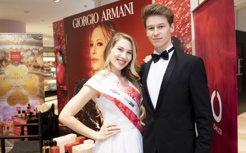 Eine Frau mit Schärpe steht lächelnd neben einem Mann im Smoking. Sie posieren vor einer Giorgio Armani-Auslage, im Vordergrund sind Kosmetikartikel zu sehen.