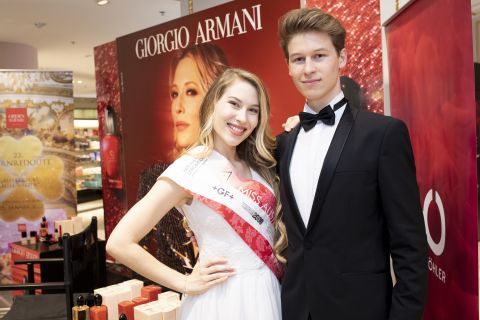 Eine Frau mit Schärpe steht lächelnd neben einem Mann im Smoking. Sie posieren vor einer Giorgio Armani-Auslage, im Vordergrund sind Kosmetikartikel zu sehen.