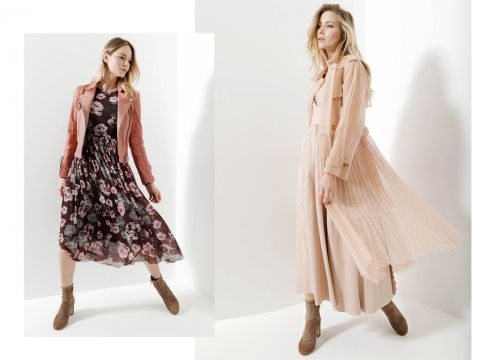 Zwei Frauen posieren in stylischen Outfits. Die linke trägt ein geblümtes Kleid mit einer rosa Jacke, die rechte ein pfirsichfarbenes Kleid mit einem passenden Mantel. Beide tragen Stiefeletten. Der helle, minimalistische Hintergrund verstärkt den modischen Fokus.