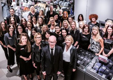 Eine Gruppe von Menschen, überwiegend Frauen, steht zusammen in einem Bekleidungsgeschäft. Sie lächeln und blicken in die Kamera, tragen eine Mischung aus schwarzer und formeller Kleidung und sind von Kleiderständern mit gefalteten Kleidungsstücken umgeben.
