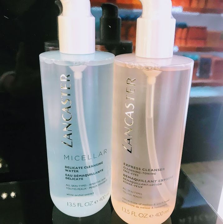Zwei Lancaster-Flaschen in einer Ladenauslage: links ein 13,5 FL OZ (400 ml) Micellar Delicate Cleansing Water und rechts ein Express Cleanser. Beide haben Pumpen und sind auf einer reflektierenden, schwarzen Oberfläche angeordnet.